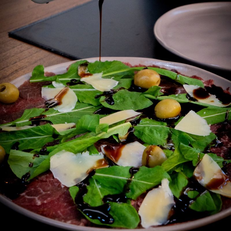 Carpaccio com rúcula, lascas de parmesão e azeitonas, recebendo calda balsâmica
