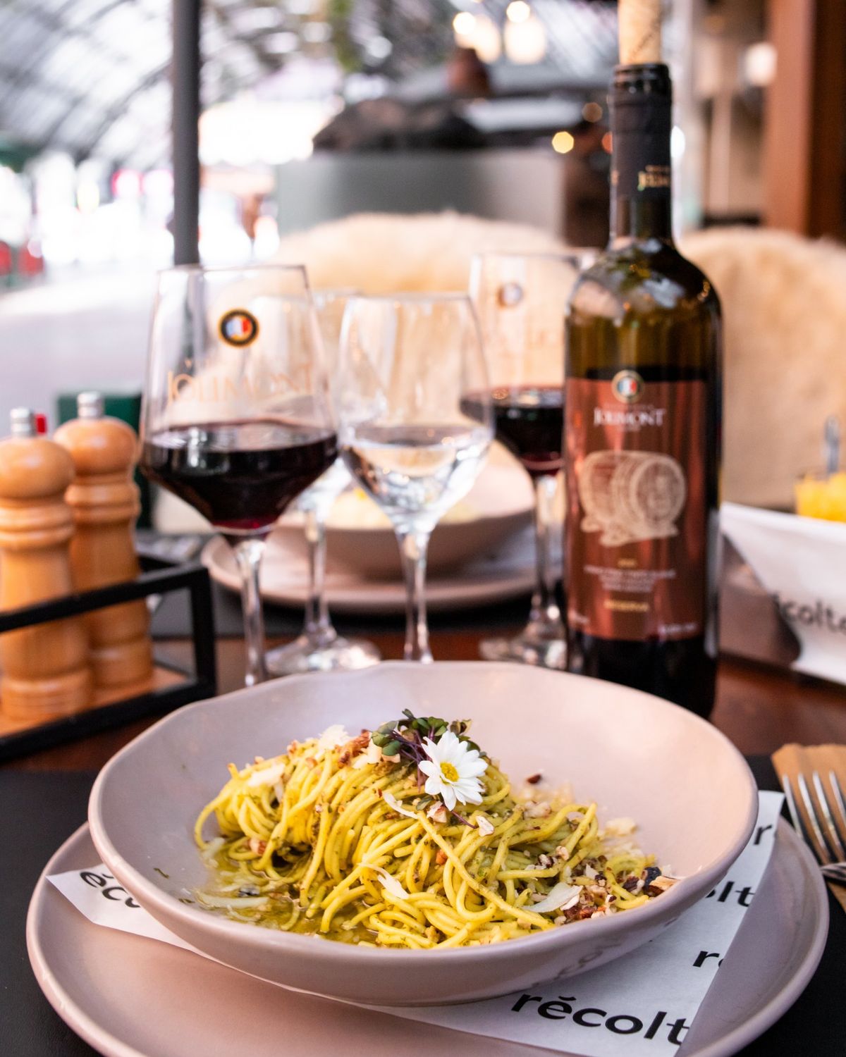 Espaguete ao pesto com flor comestível, taças de vinho tinto e uma garrafa Jolimont na mesa do Récolte, sob a cobertura da Rua Coberta