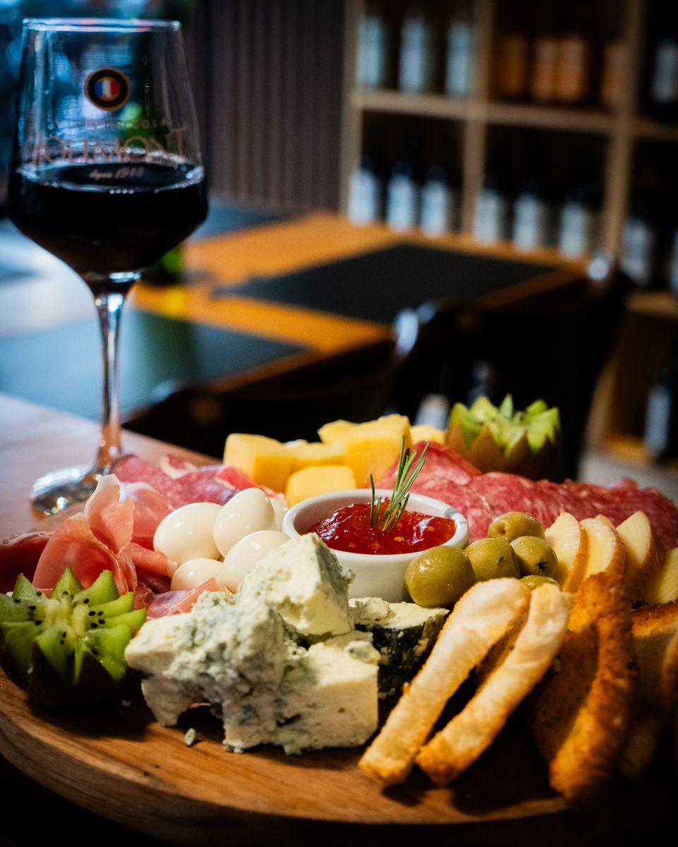 Tábua de frios com parma, salame, queijos, gorgonzola, ovos de codorna, azeitonas e torradinhas, ao lado de uma taça de vinho tinto