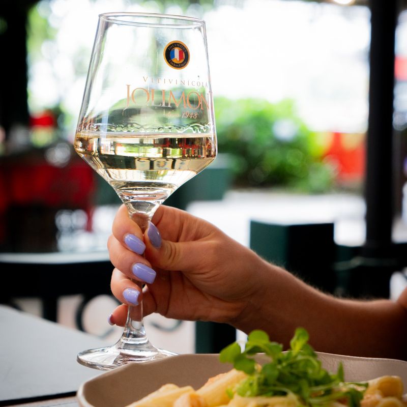 Mão segura uma taça de vinho branco diante de um prato de massa, com o verde da Rua Coberta ao fundo